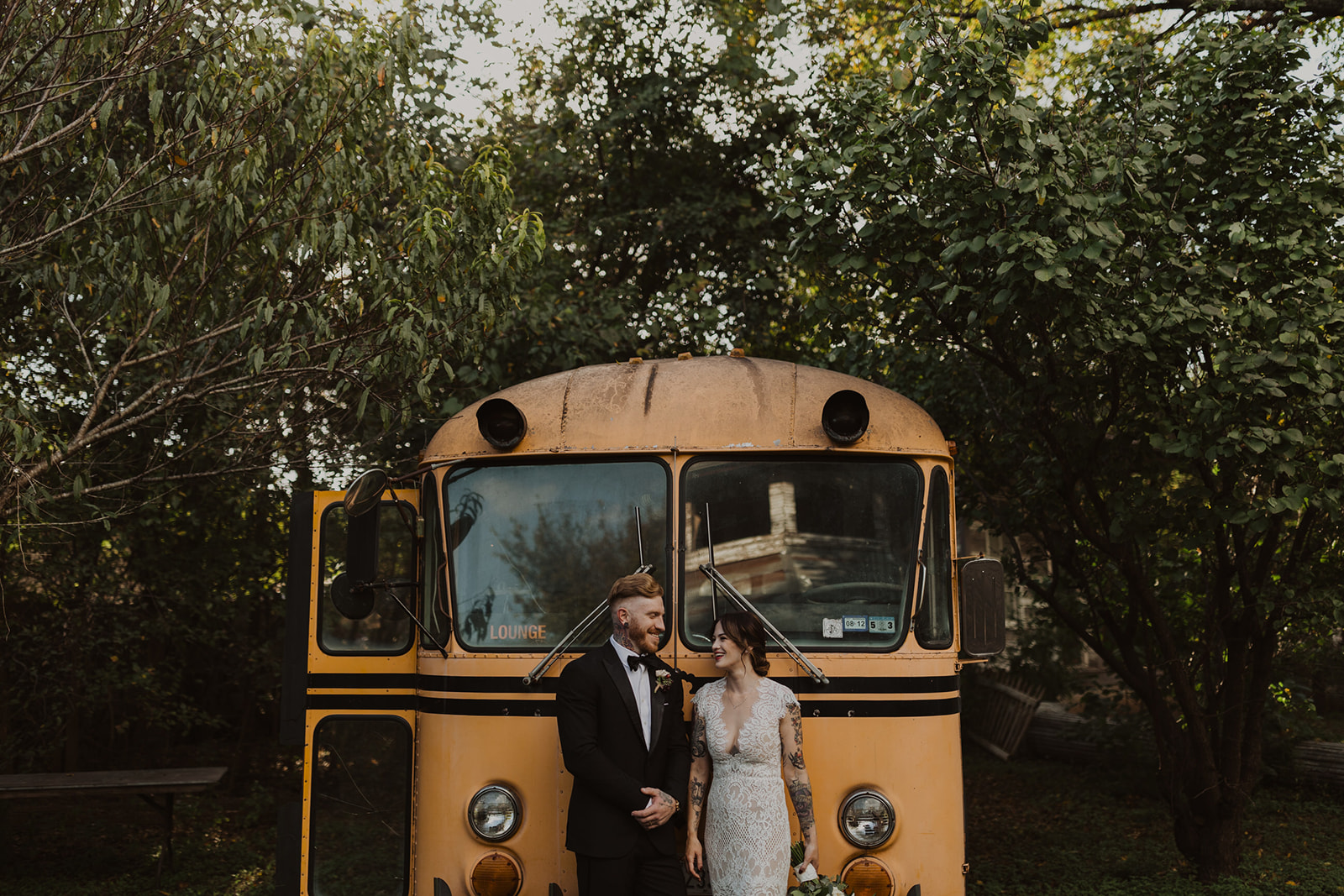 couple poses beside school bus at outdoor wedding