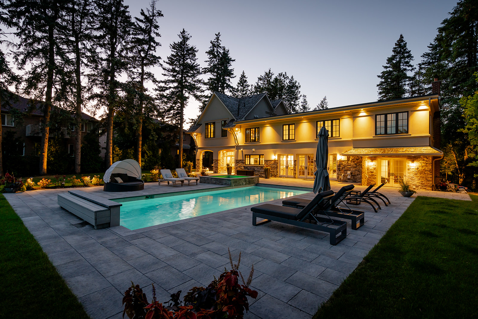 A backyard with an inground pool with furniture on either side and the house with lights on in the background.