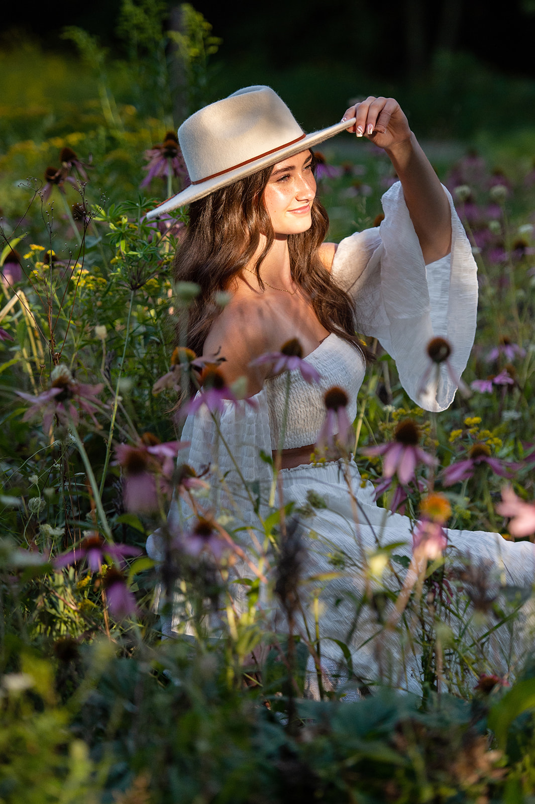 Beautiful brown-haired girl sitting in wildflowers, holding the rim of her hat, looking into the distance. 