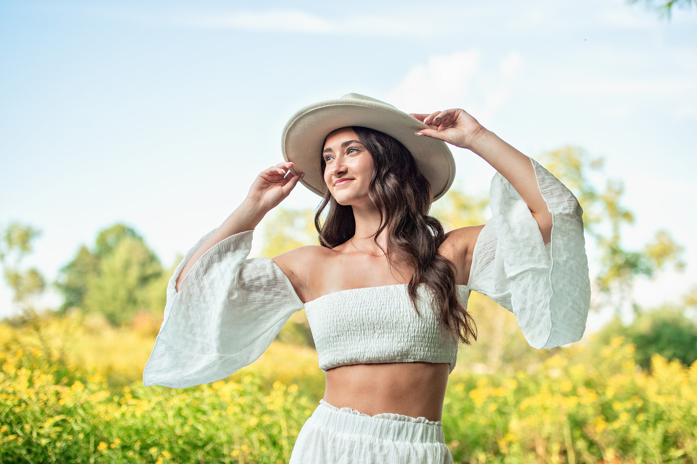 Beautiful high school senior girl in a white flowy dress, holding her hat and looking into the distance.