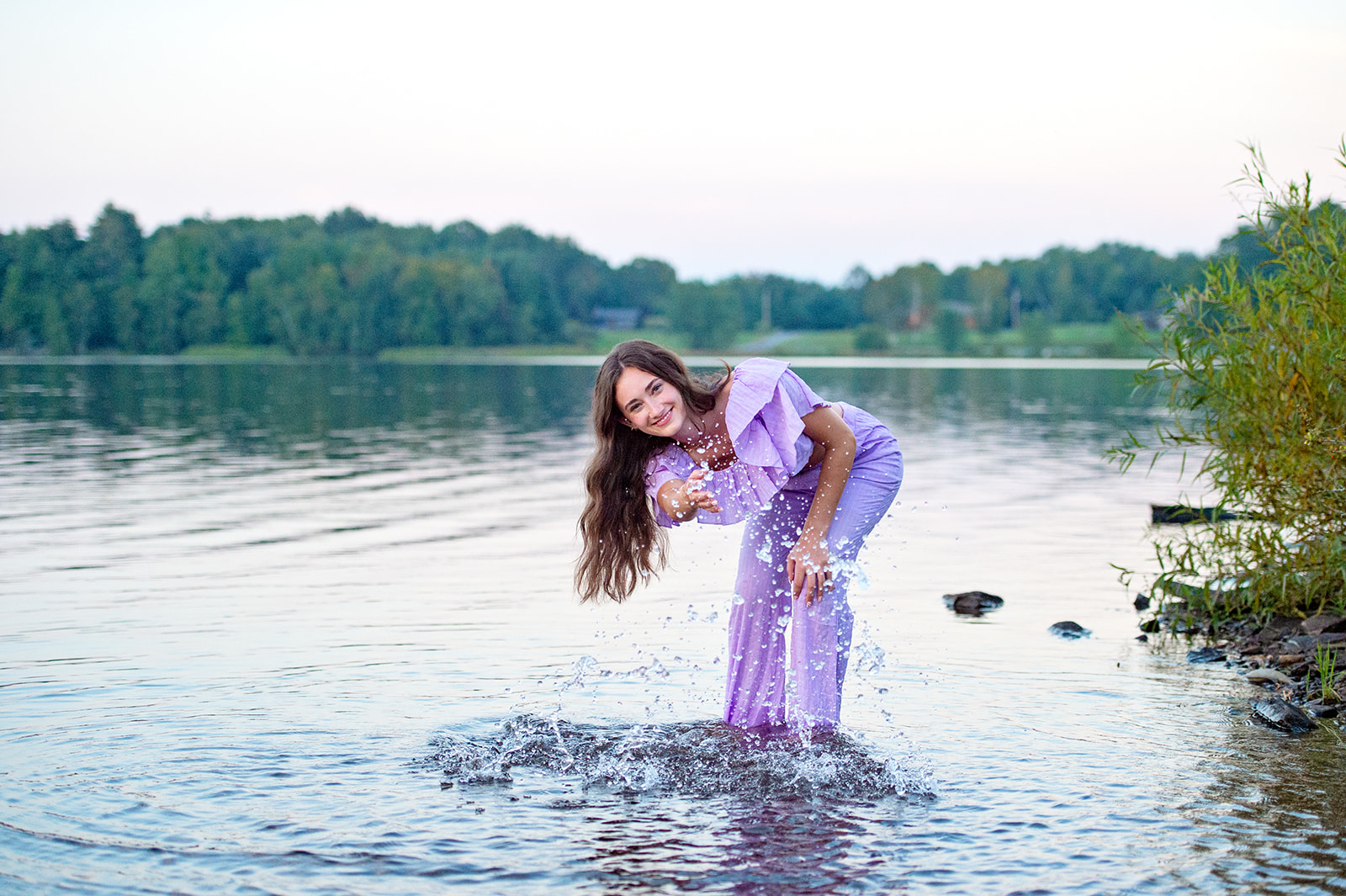 A high school senior in a lake, splashing water toward the photographer and laughing. 