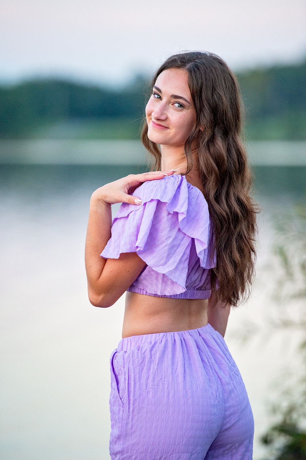 A senior girl standing in the lake in a purple two piece outfit looking over her shoulder with a subtle smile. 