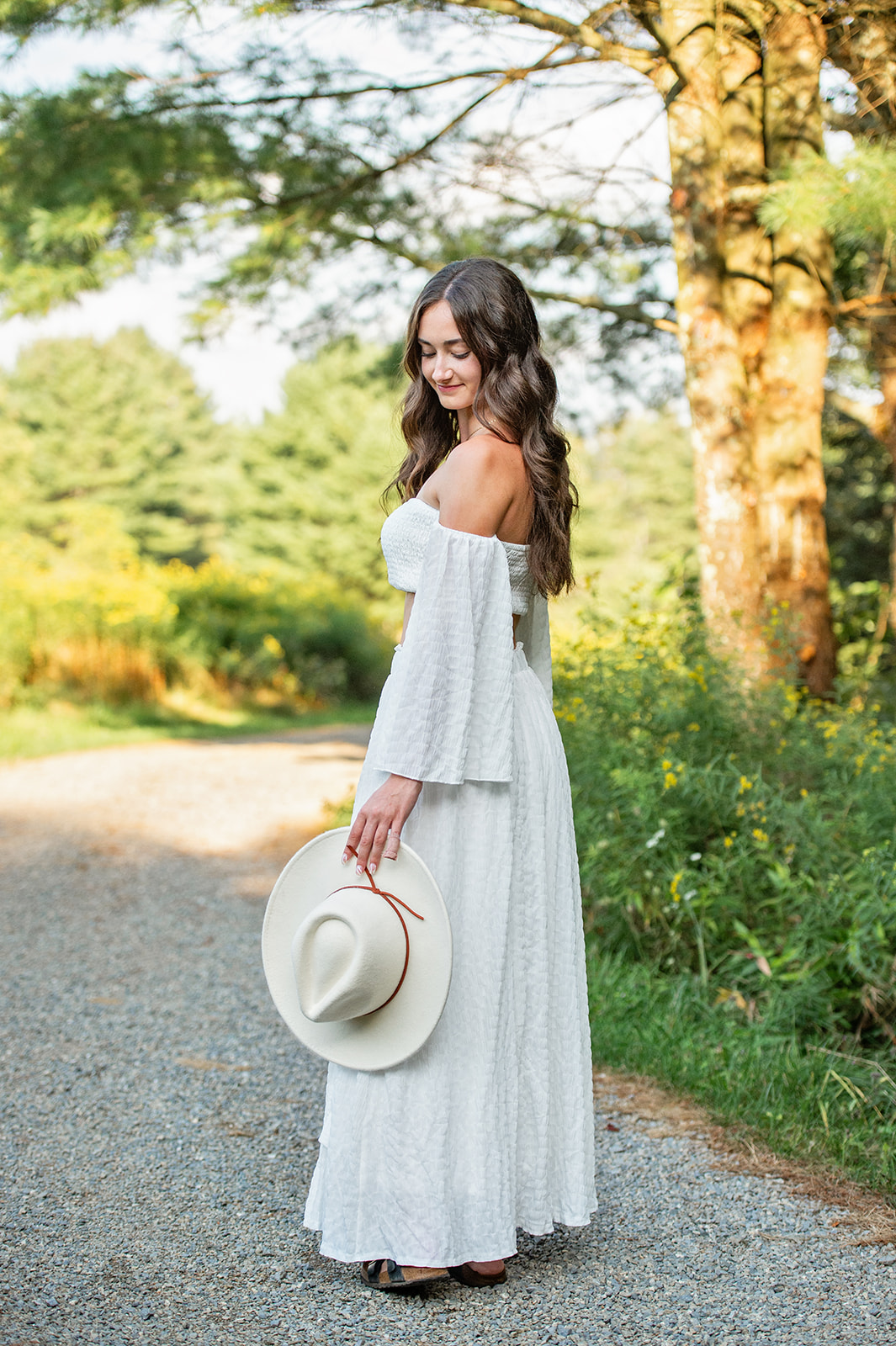 Senior girl in a two piece white dress holding a white hat, looking down at it. 