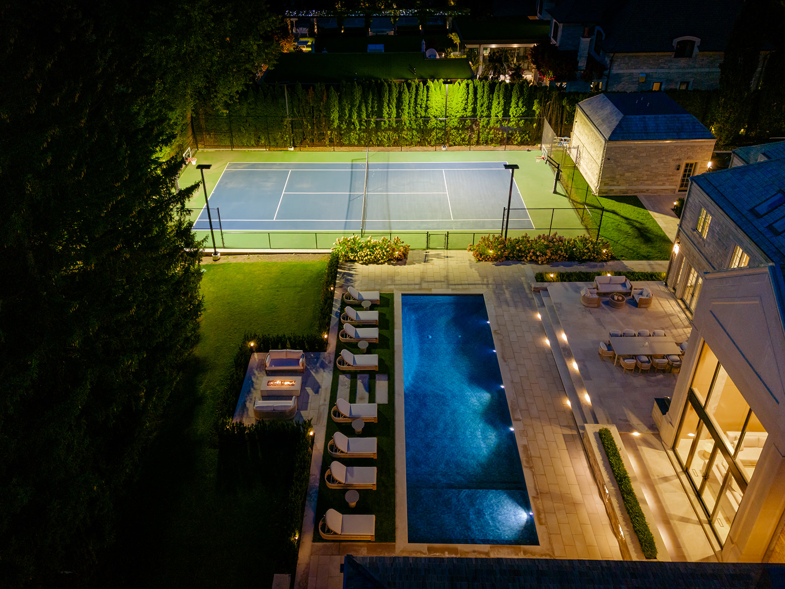 A done-shot of the backyard with the inground pool and tennis court lit up.