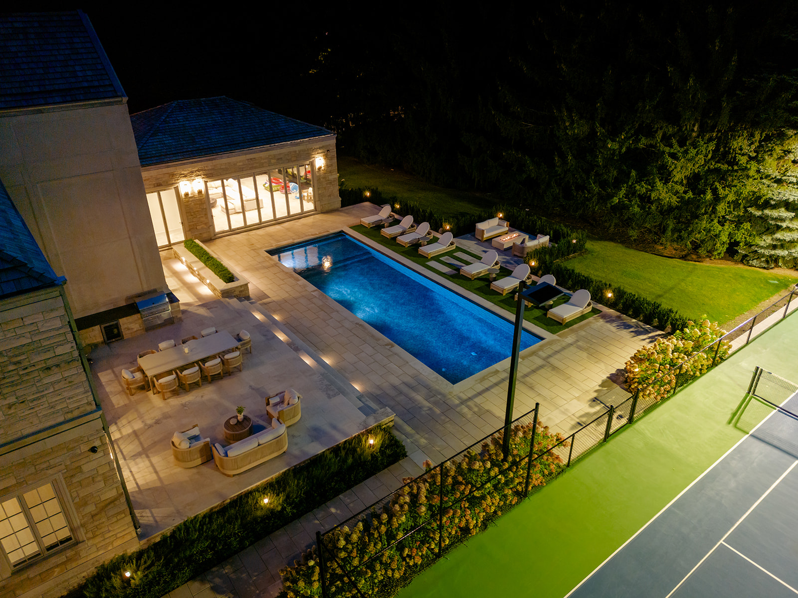 A done-shot of the backyard with the inground pool and tennis court lit up.