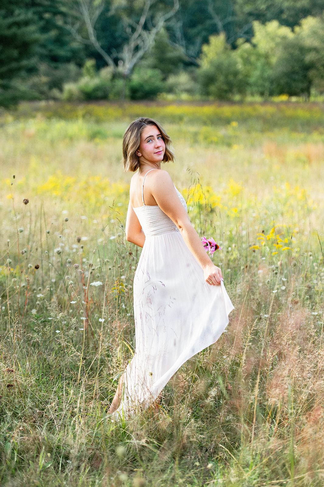 Beautiful senior girl walking through a wildflower field with a flowing dress, looking back smiling. 