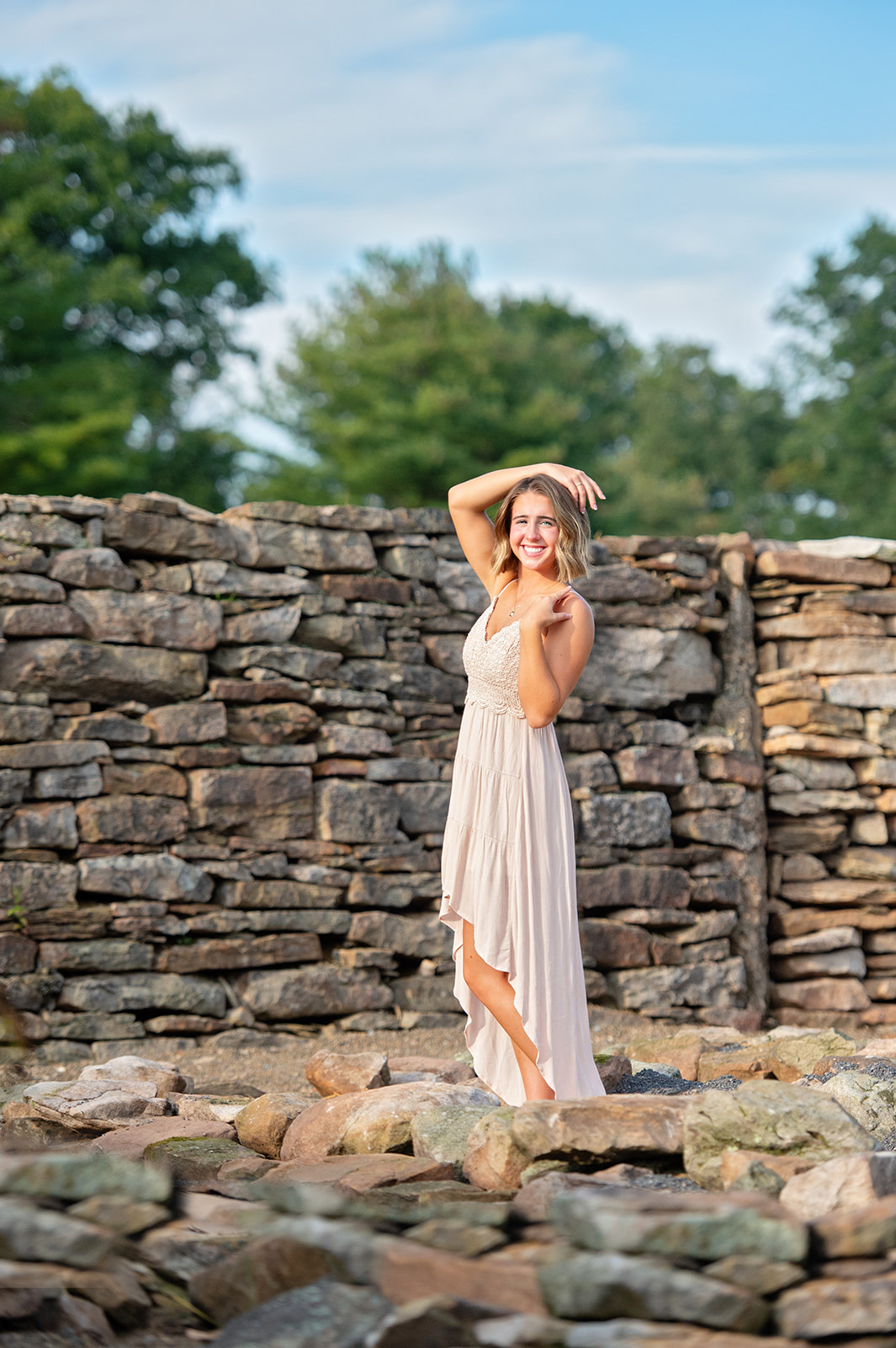 Gorgeous high school senior posing next to a stone wall in a field with the sun illuminating her. 