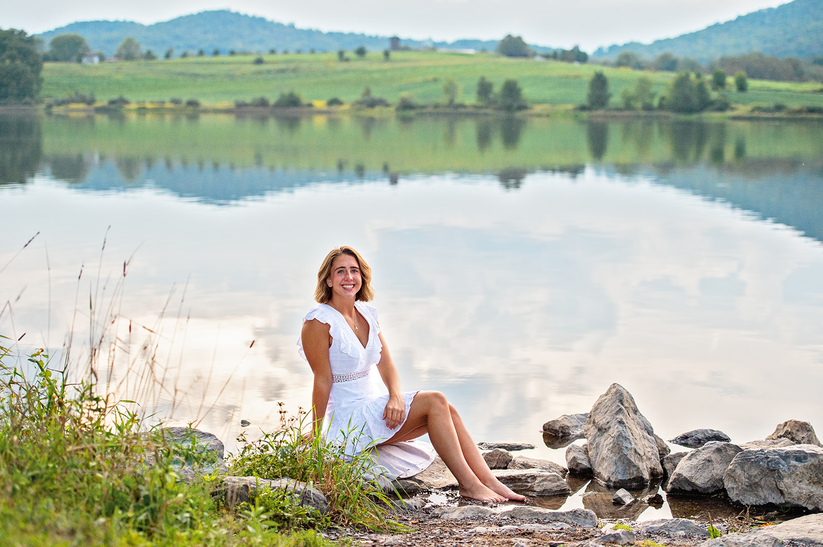 High school senior girl in white sundress sitting on rocks by the lake at sunset. 