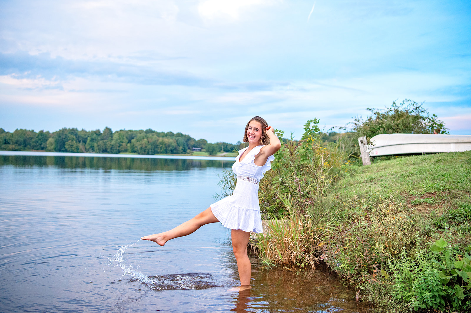 Senior girl in a white sundress kicking water in lake laughing. 