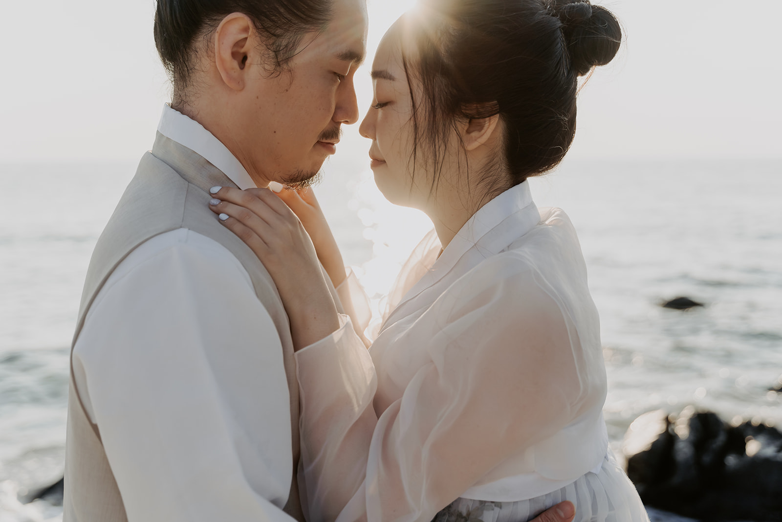 A couple in traditional Korean clothing standing by the ocean almost kissing for their pre-wedding