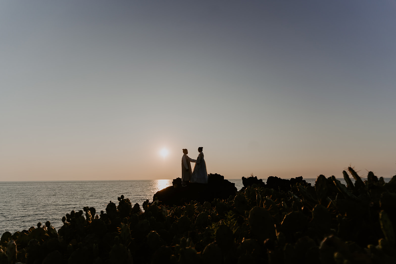 A pre-wedding couple standing while holding hands on a rock overlooking the ocean in Korea during sunset.