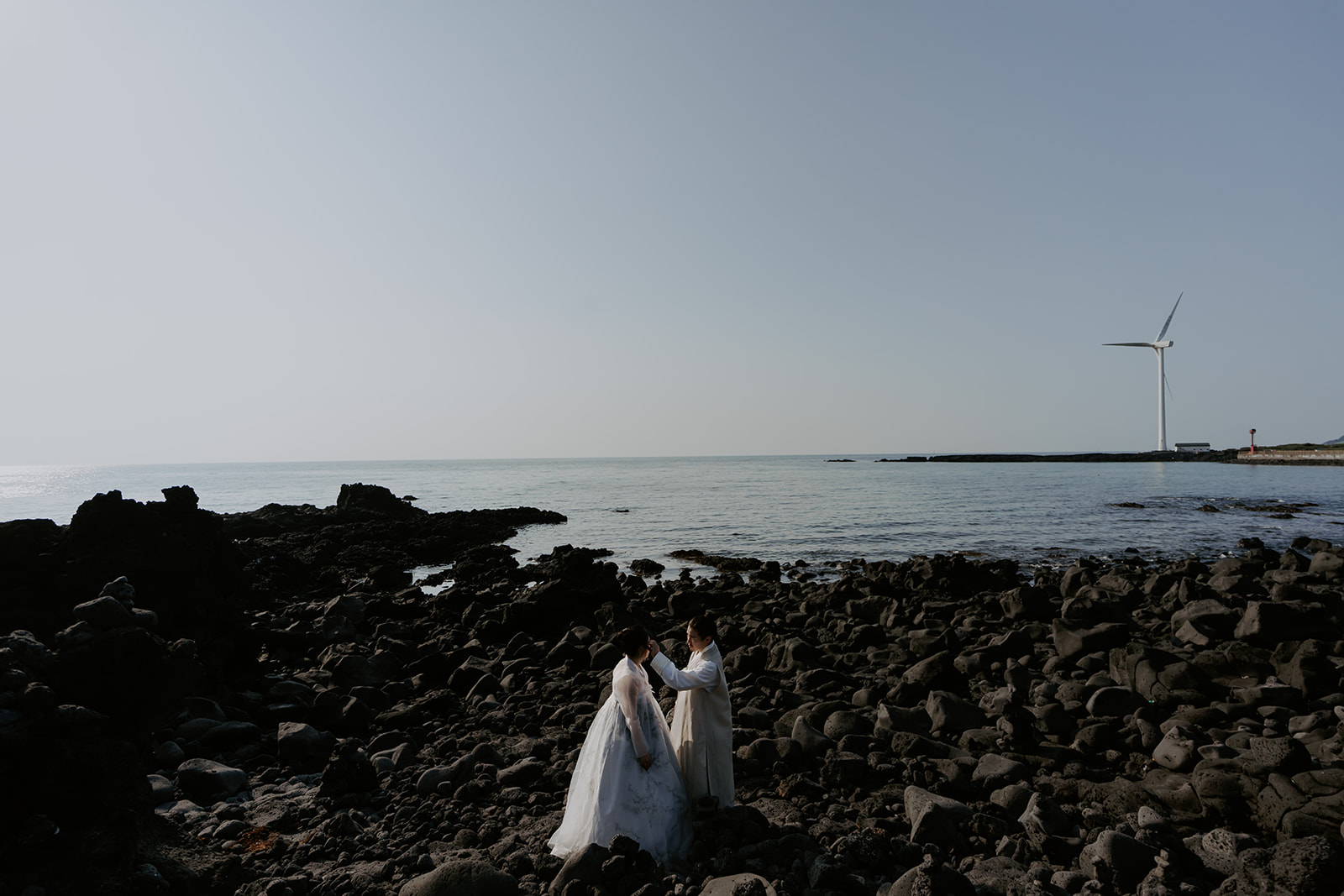 A pre-wedding couple standing on rocks in front of a wind turbine in Korea.