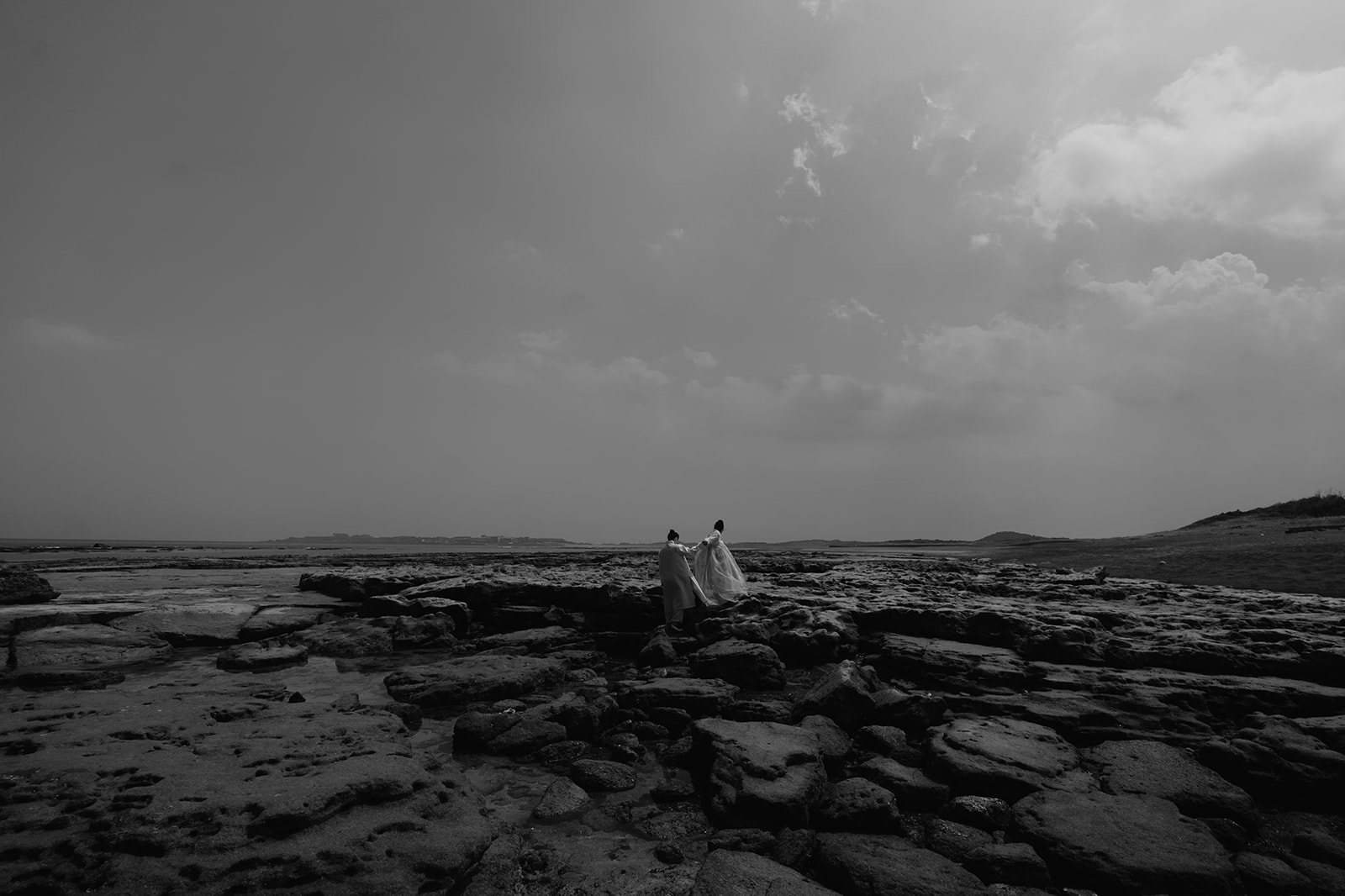 A pre-wedding photo of a couple walking on a rocky beach in Korea, captured in black and white.