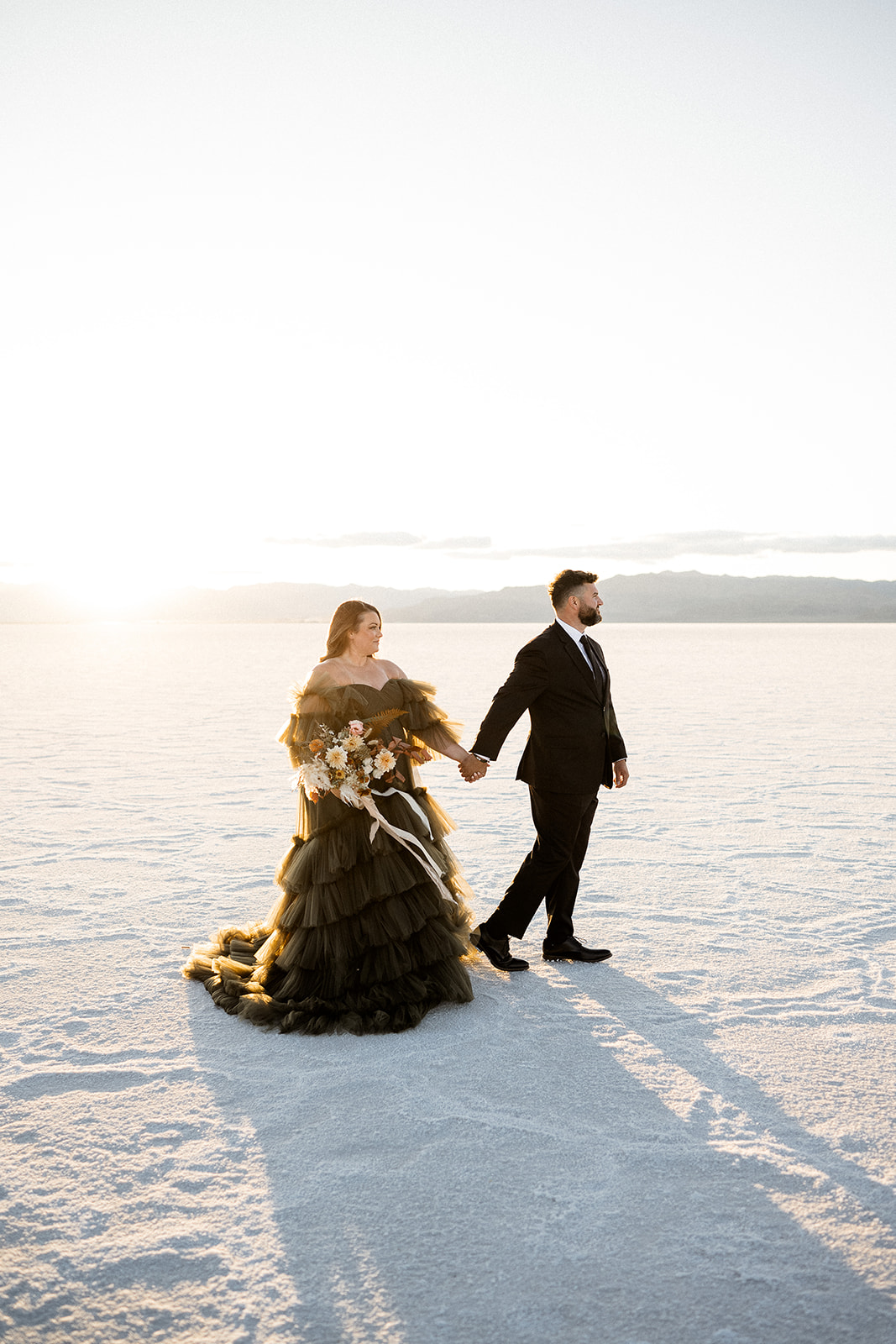 Couple walking hand in hand at sunset on the Salt Flats, casting long shadows, in formal attire