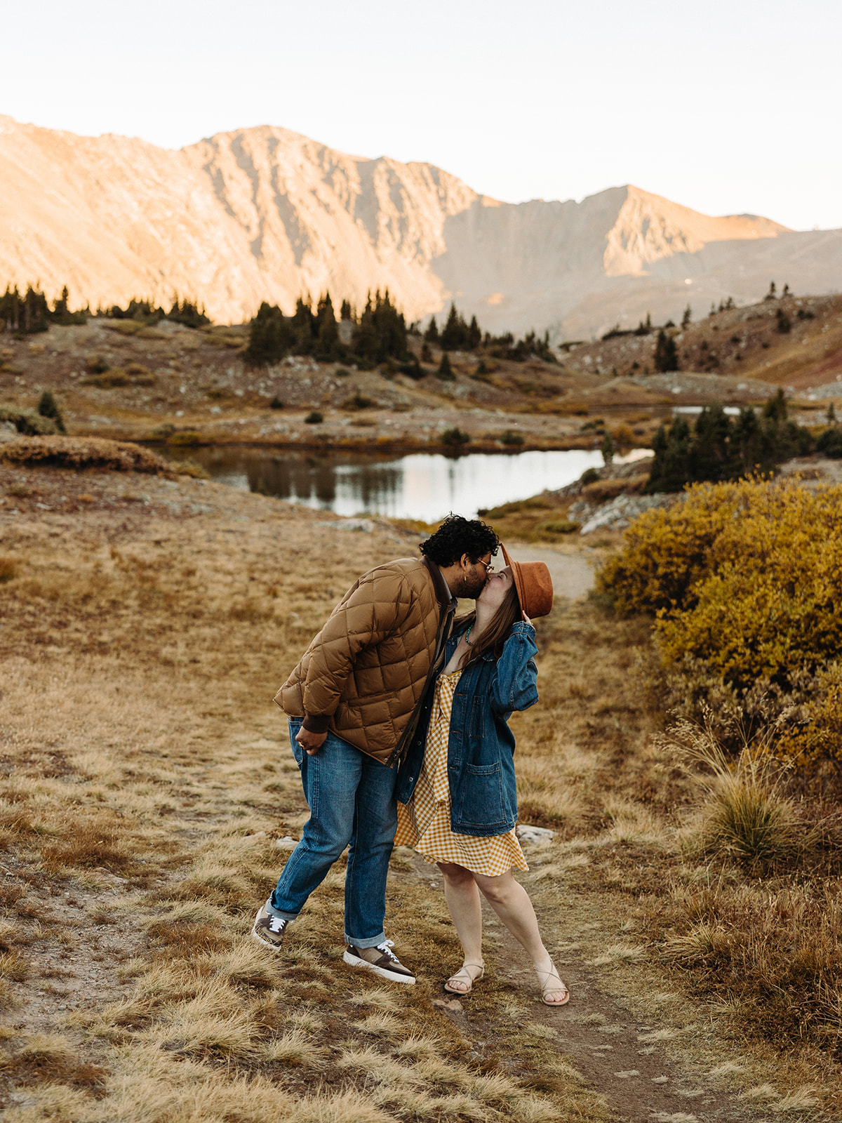 Woman in a yellow dress, brown hat, and jean jacket holds her hat as she looks up to kiss her fiance near the lake.