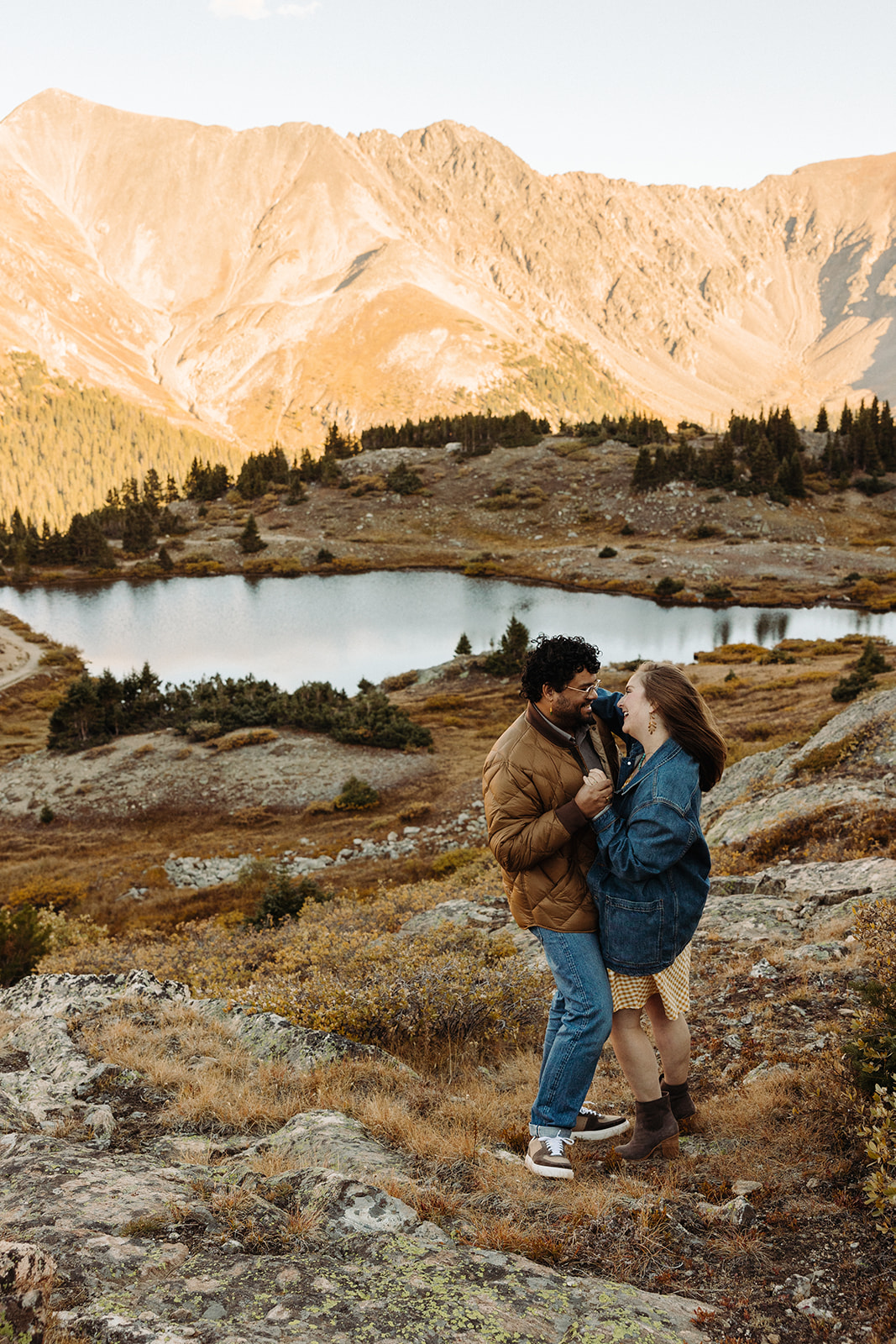The man, in his brown puffy jacket and blue jeans, holds his fiance in the grassy valley of the towering mountains.