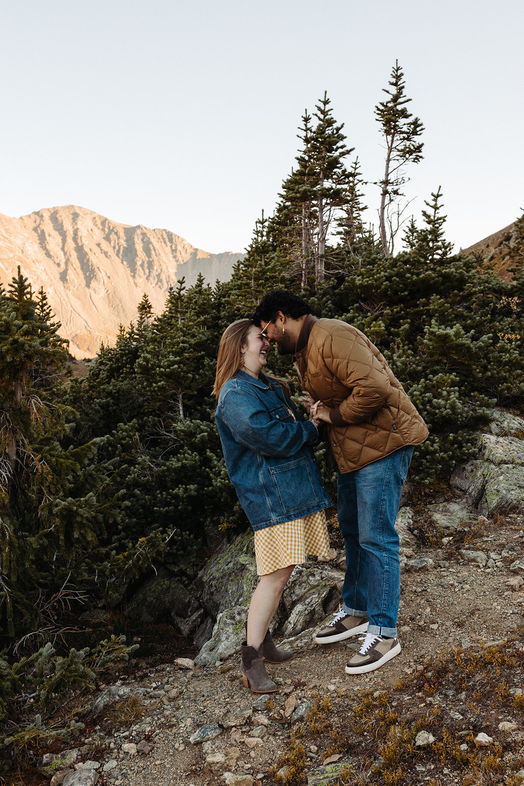 The couple smile and laugh while touching foreheads amongst the spruce trees and rocky trails.