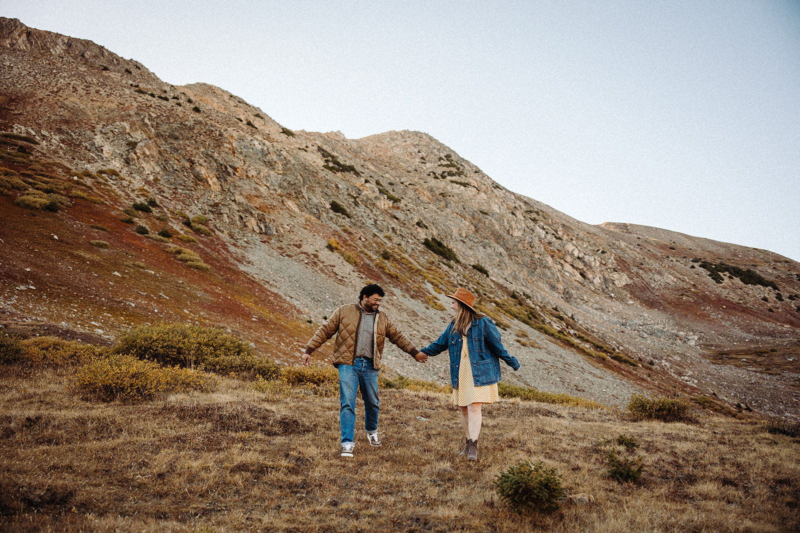 Man and his fiance holding hands as they walk through a grassy meadow along the rusty hills.