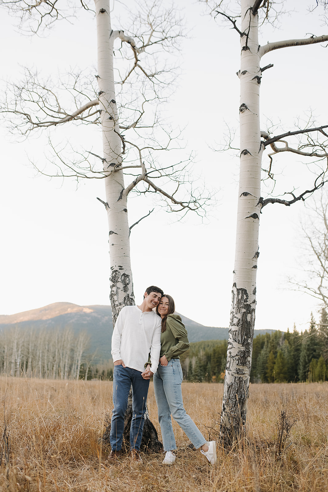 Newly engaged couple standing against a backdrop of Colorado Aspen trees smiling at the camera for a photo