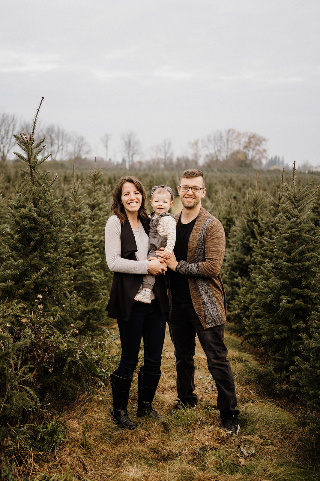 A family of three standing outside with the Christmas Trees.