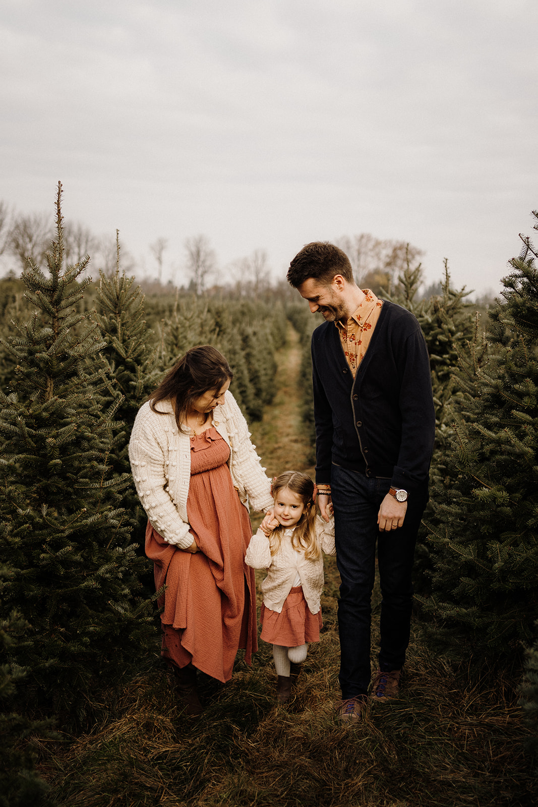 Parents holding their child's hands, walking down a path between Christmas Trees.