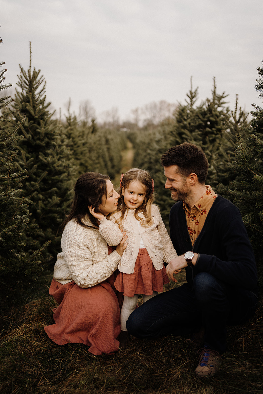 Parents kneeling with their child between Christmas Trees outside.