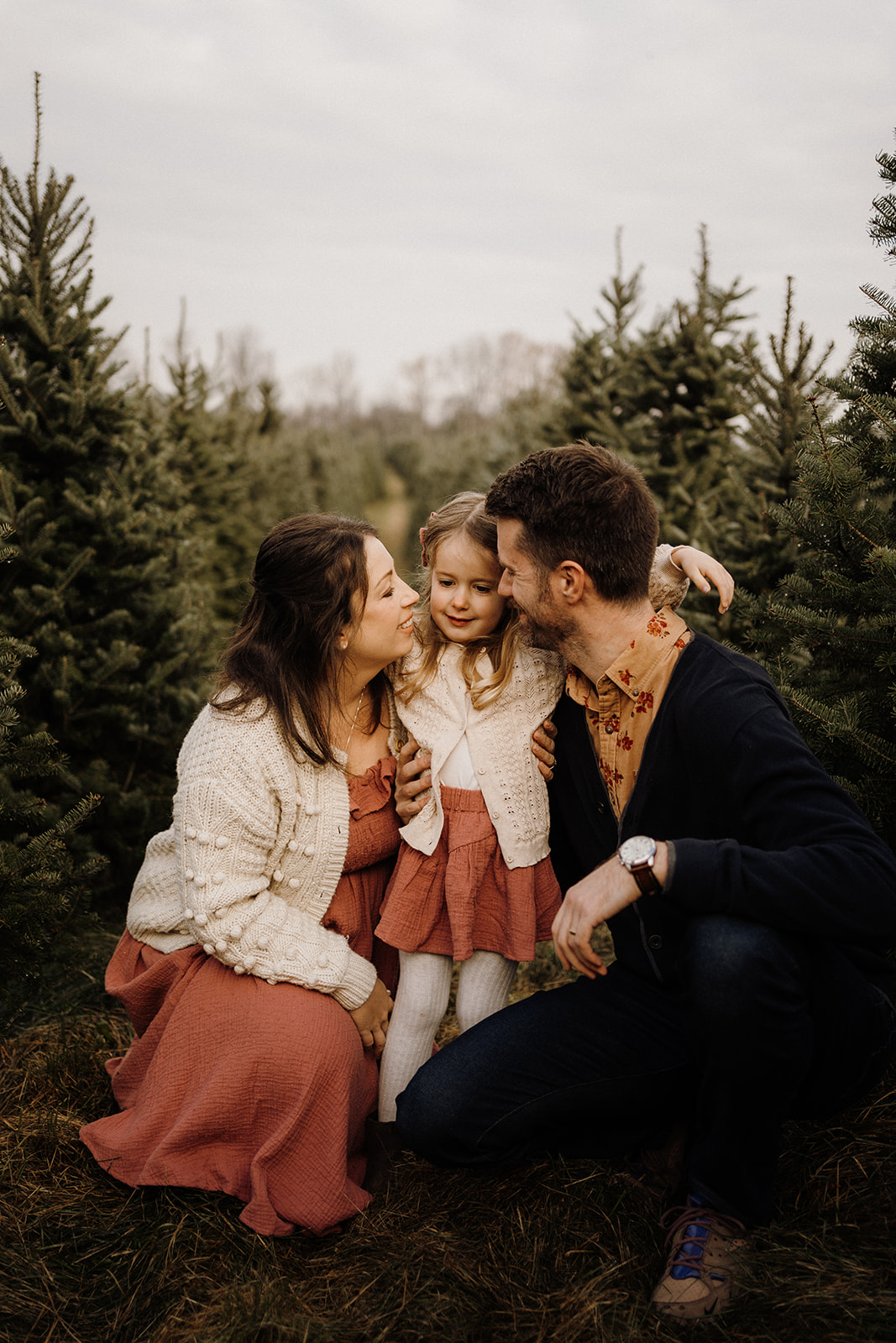 Parents kneeling with their child between Christmas Trees outside.