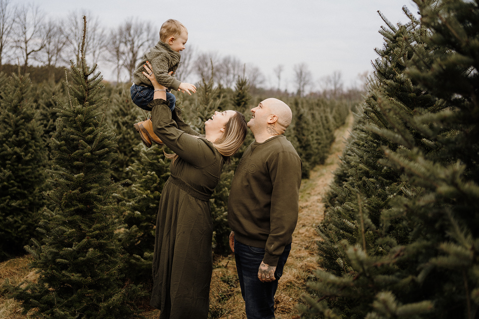 A family of three standing outside surrounded by Christmas Trees.