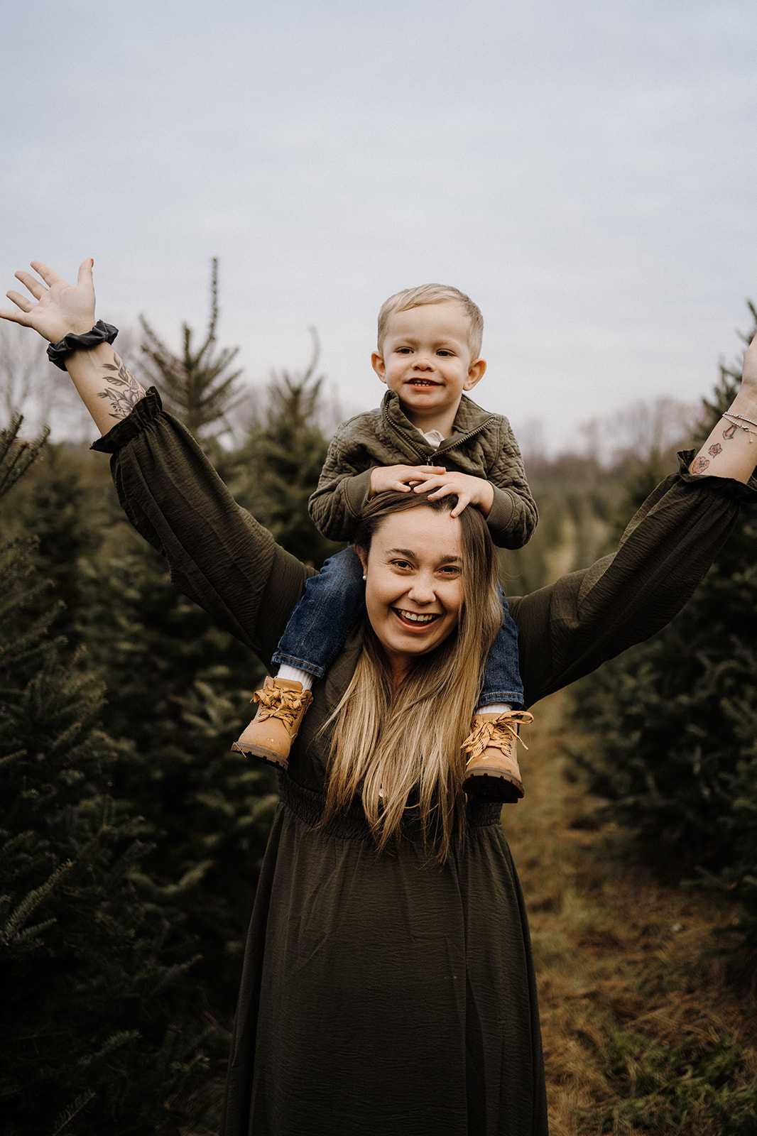 A mother with her son on her shoulders outside between Christmas Trees.