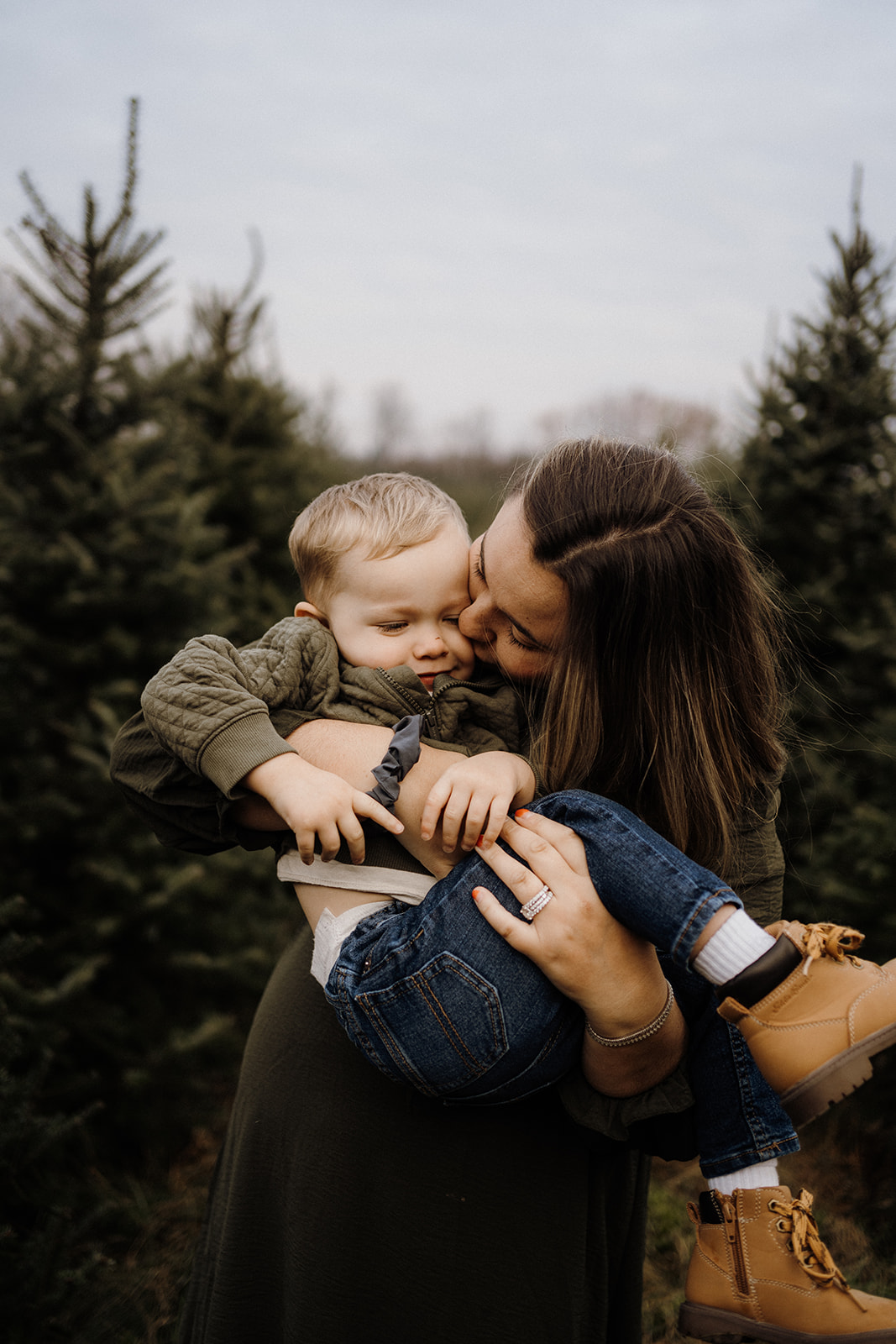 A mother kissing her son on the cheek in her arms outside between Christmas Trees.