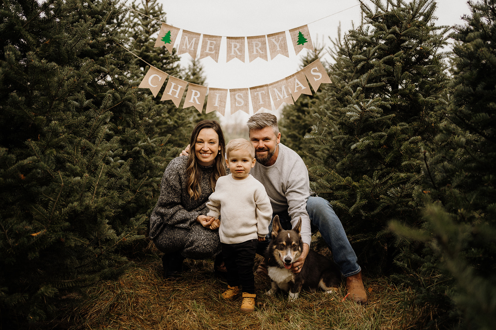 A family of three sitting underneath a 'Merry Christmas' banner surrounded by Christmas Trees.