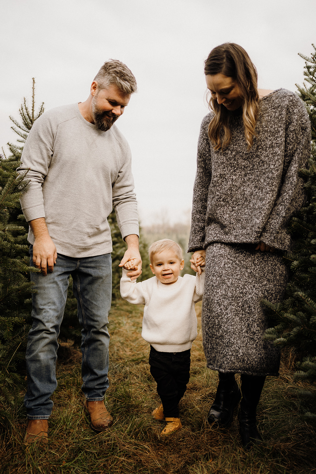 Parents holding their child's hands while walking outside between Christmas Trees.