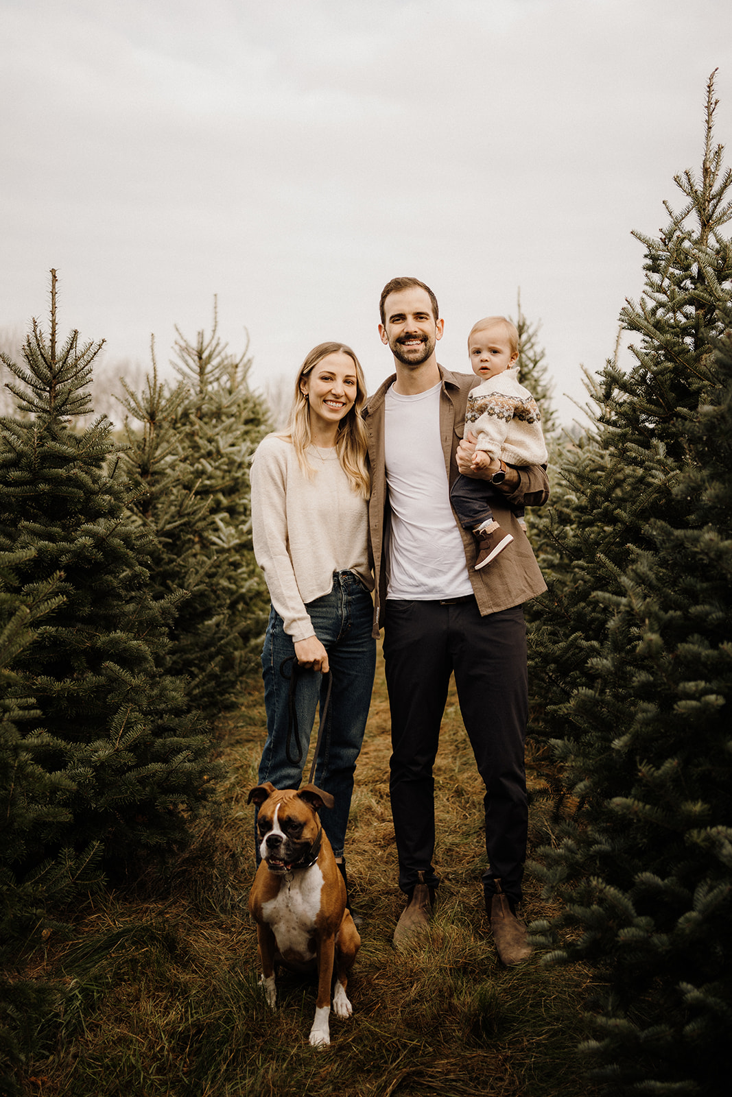 A family of three standing with Christmas Trees behind them.