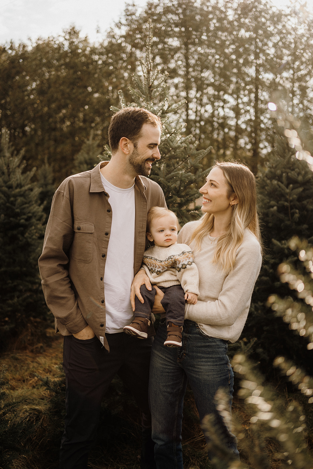 A family of three standing surrounded by Christmas Trees.