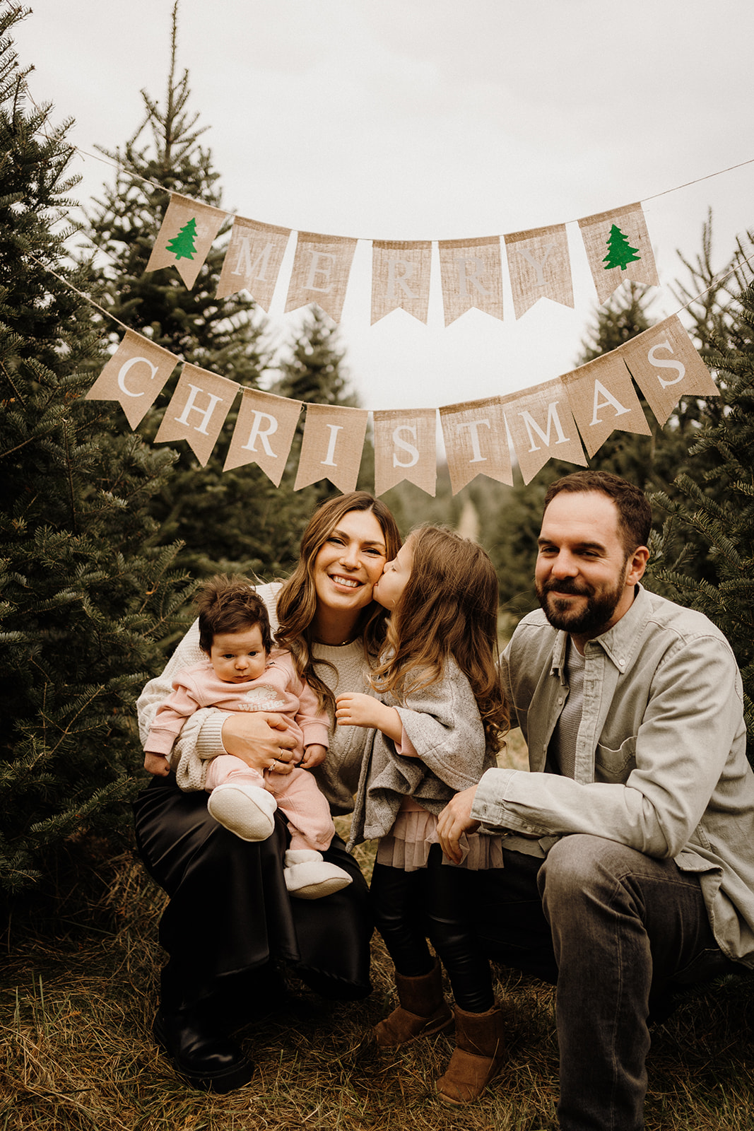 A family of four sitting underneath a 'Merry Christmas' Banner with Christmas Trees beside them.