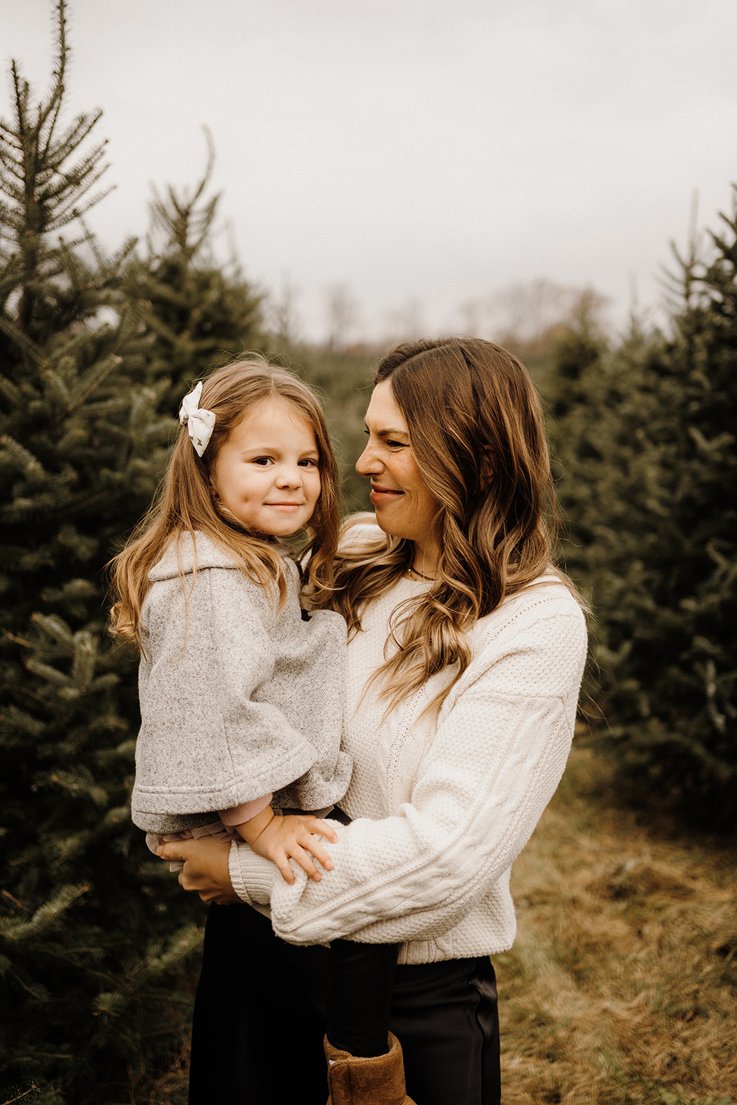 A mother holding her daughter in her arms in front of Christmas Trees.