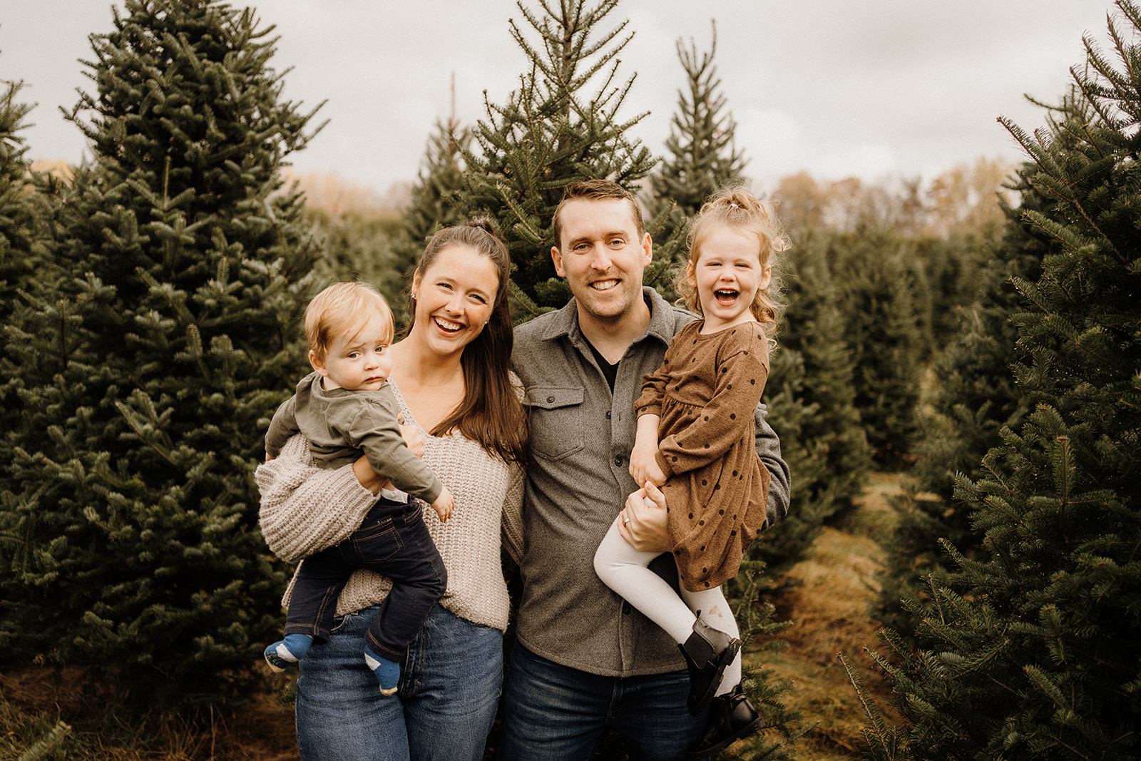 A family of four standing outside in front of Christmas Trees.