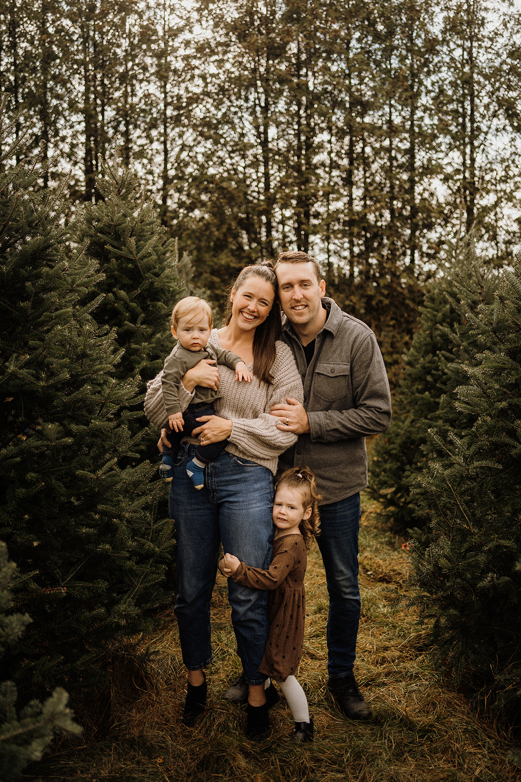 A family of four standing outside in front of Christmas Trees.