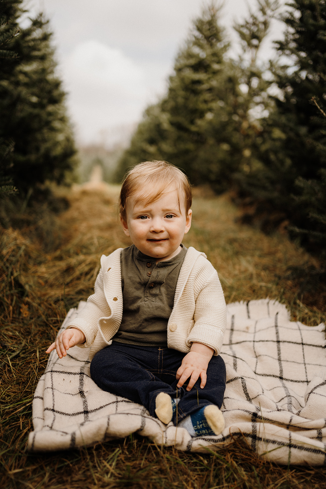 A little boy sitting on a blanket outside between Christmas Trees.