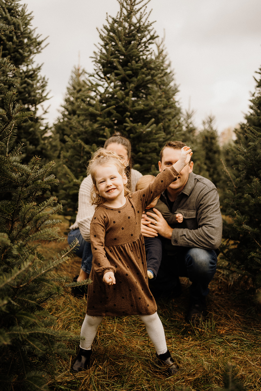 A little girl standing in front of her parents outside between Christmas Trees.