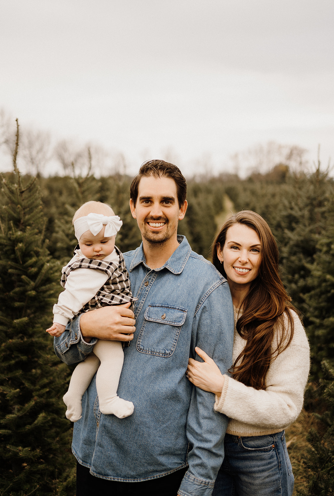 A family of three standing outside in front of Christmas Trees.