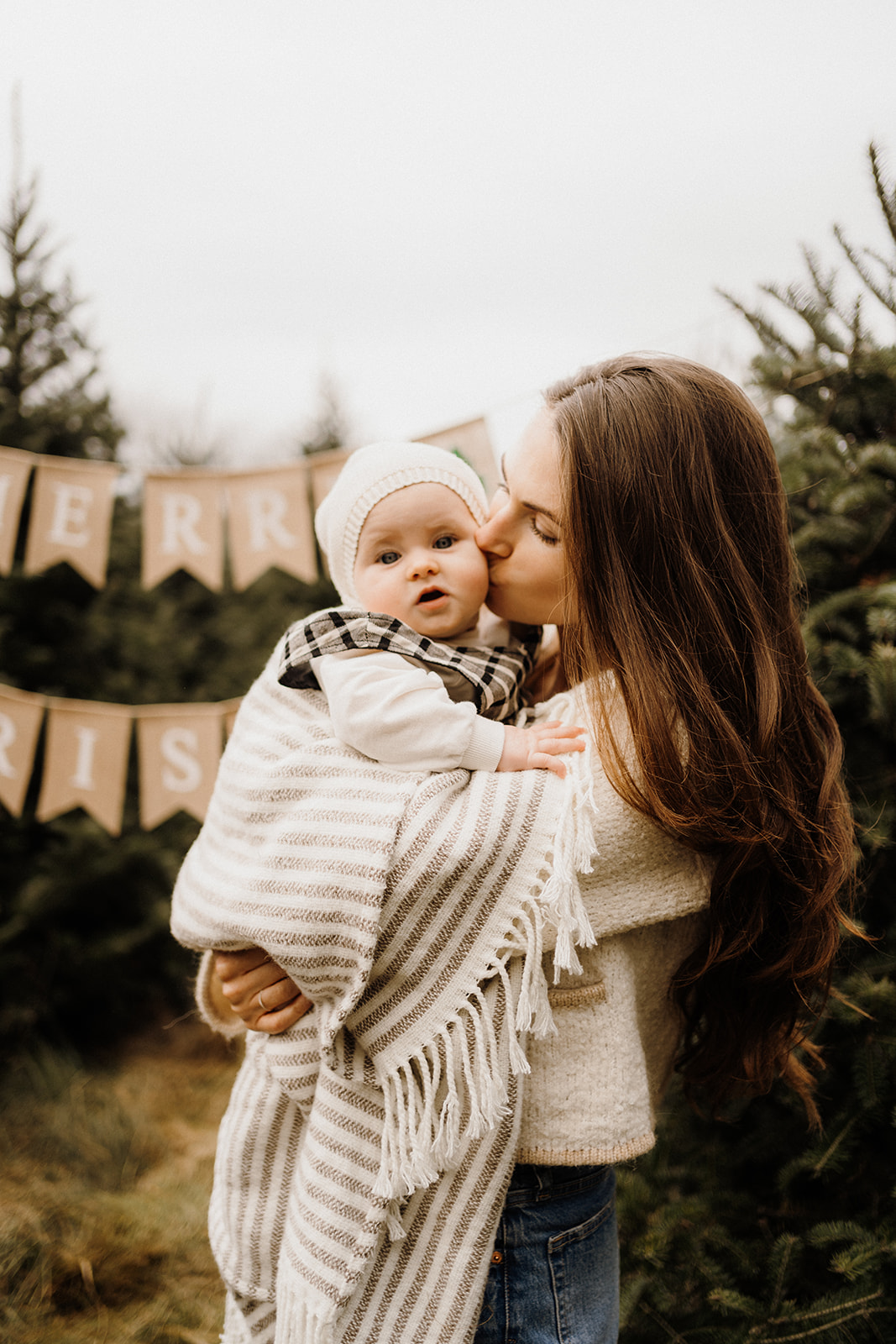 A little girl being held by her mother outside in front of Christmas Trees.