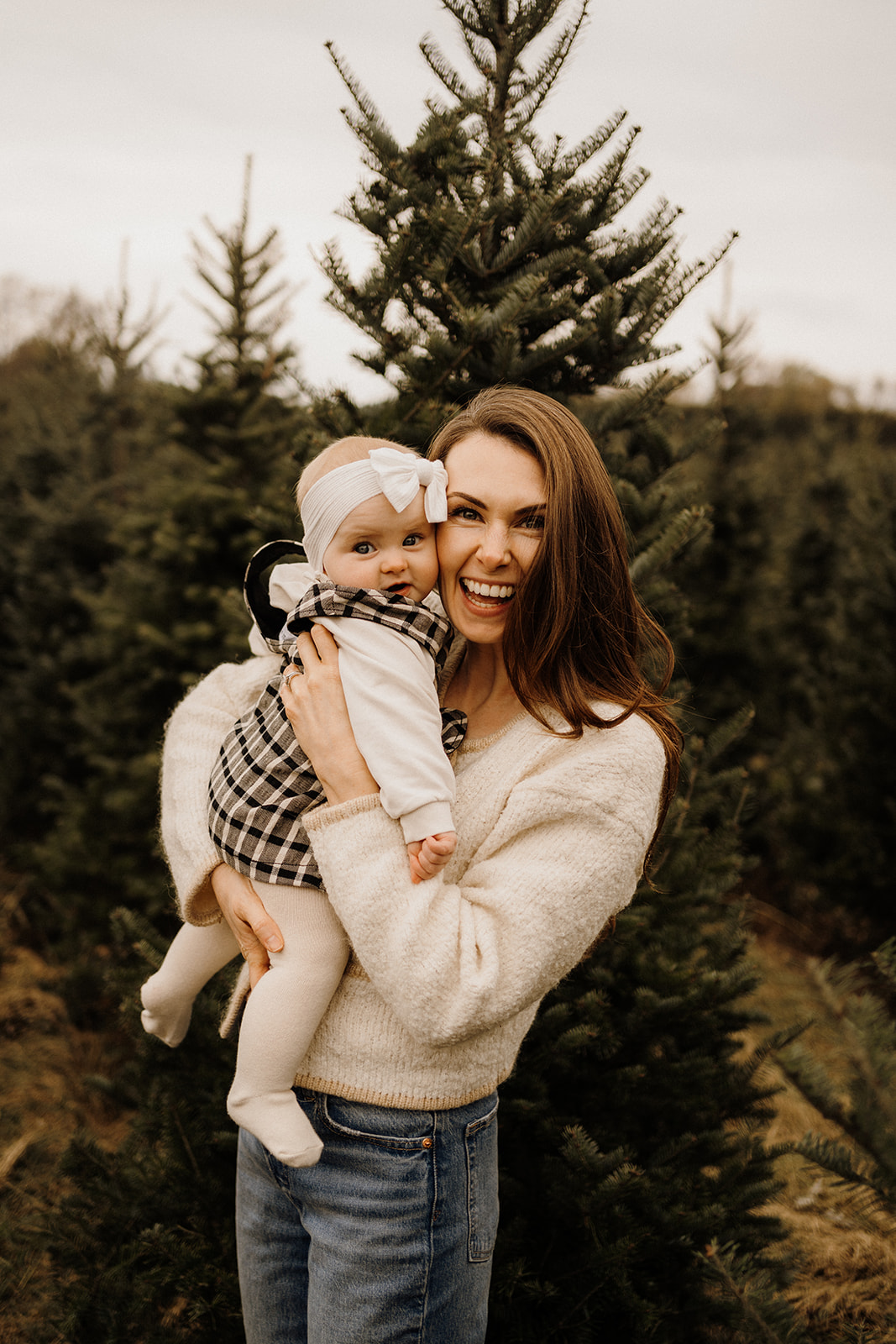 A mother holding her daughter in her arms in front of Christmas Trees outside.