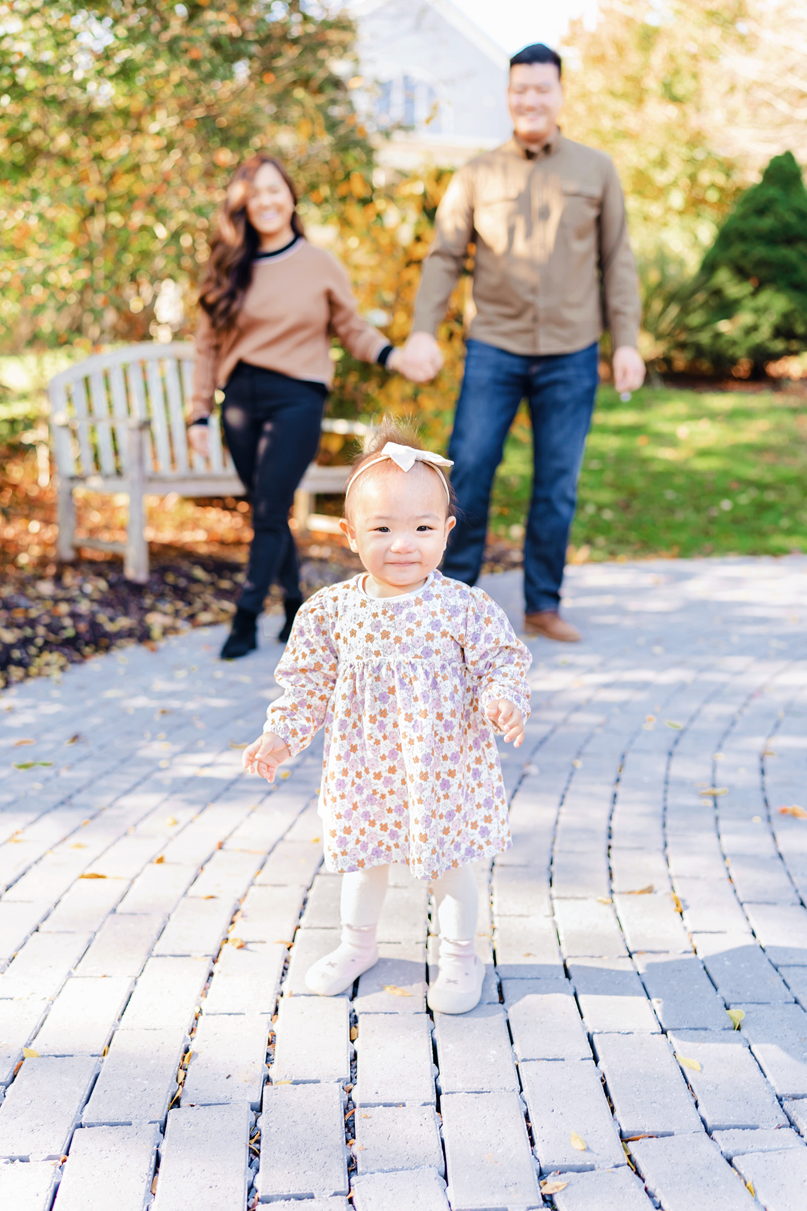 Baby stands in front of her parents smiling during her fall photo session at the Howard County Conservancy