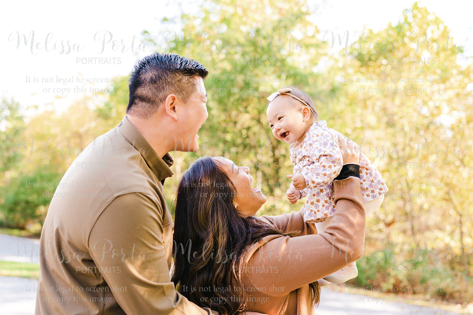 Family smiles at their baby as she laughs at them with fall foliage as a backdrop in Howard County, MD