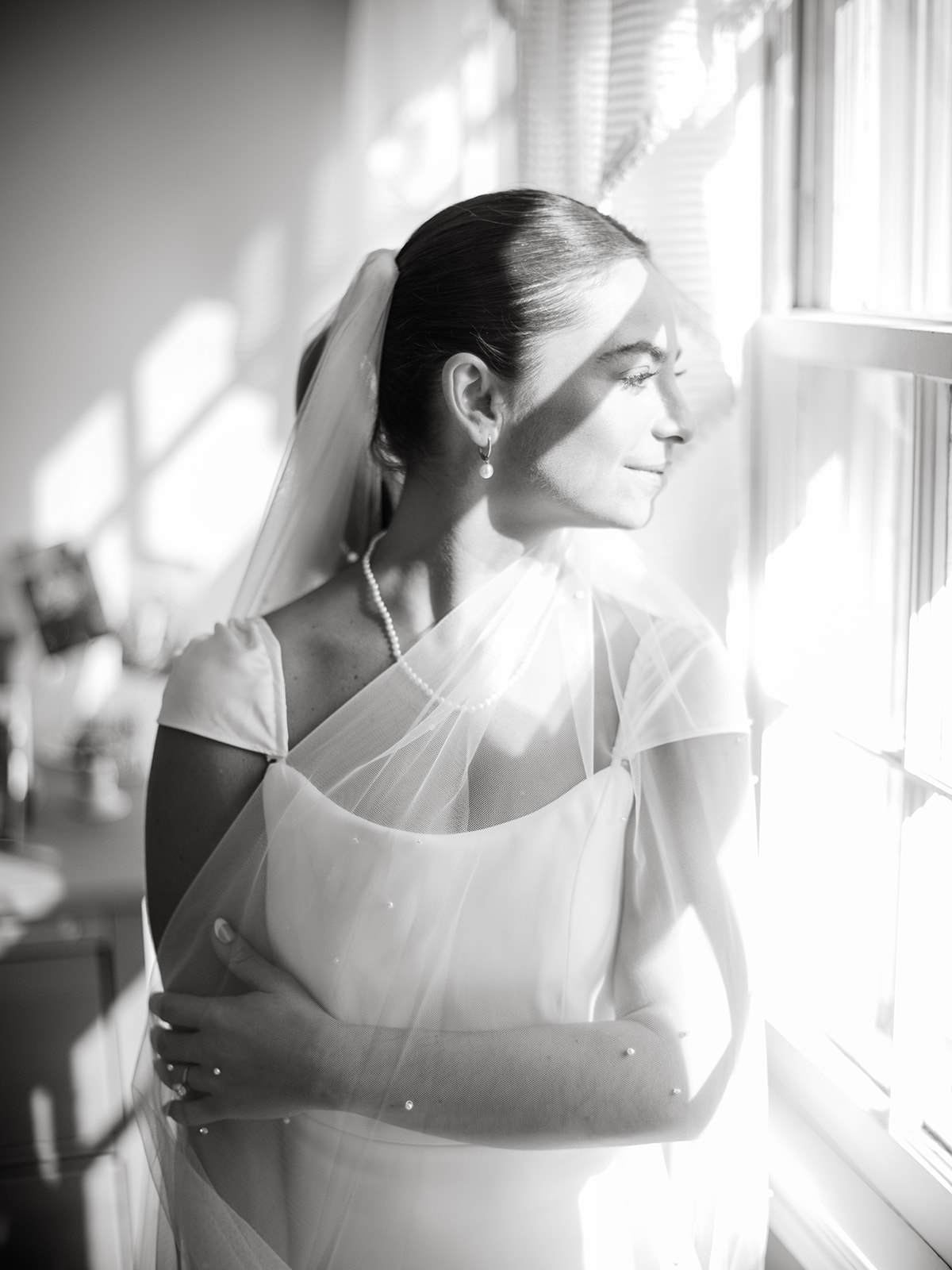 A bride looks out the window after getting ready for her wedding day, letting the light shine on her face.