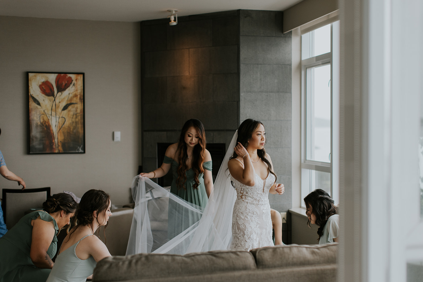 a bride looks out the window while her bridesmaids fix up her dress 