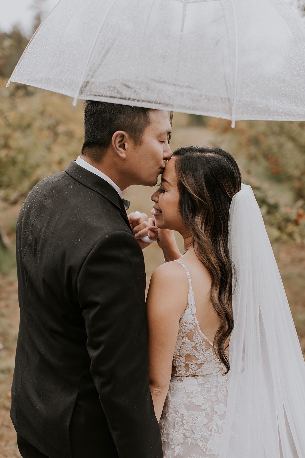a groom kisses the forehead of his bride while they hold an umbrella