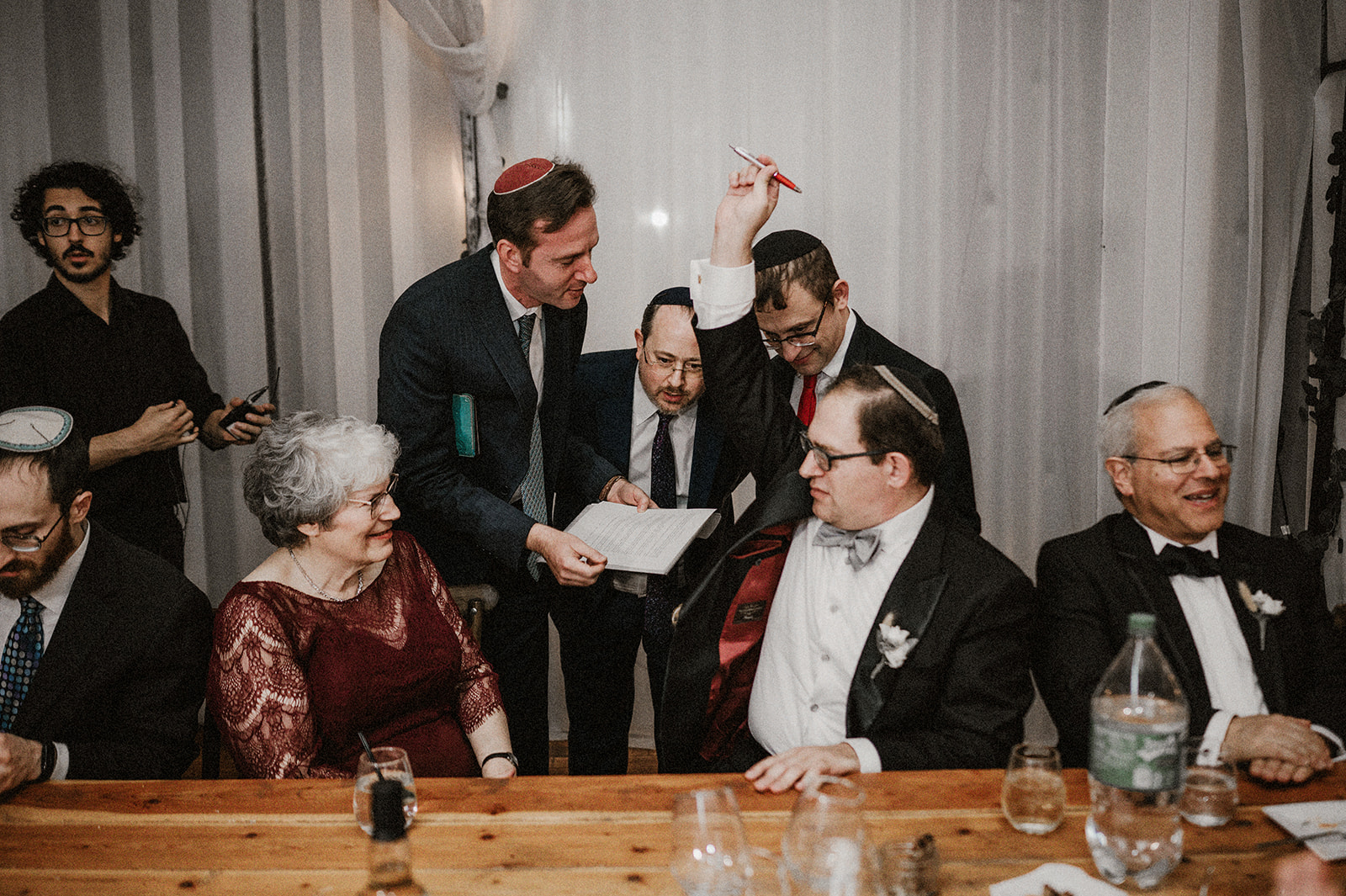 groom joined by family and friends during khasan's tisch as he raises pen to signify signature in modern orthodox jewish
