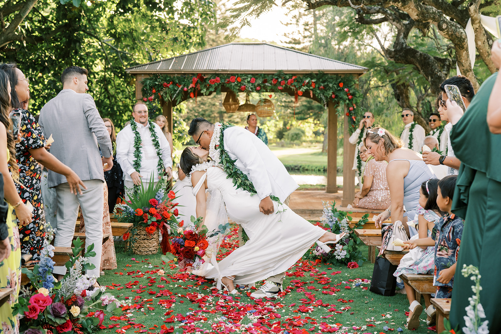 A couple at their outdoor wedding ceremony sharing a kiss with guests lined up along a flower-strewn aisle.